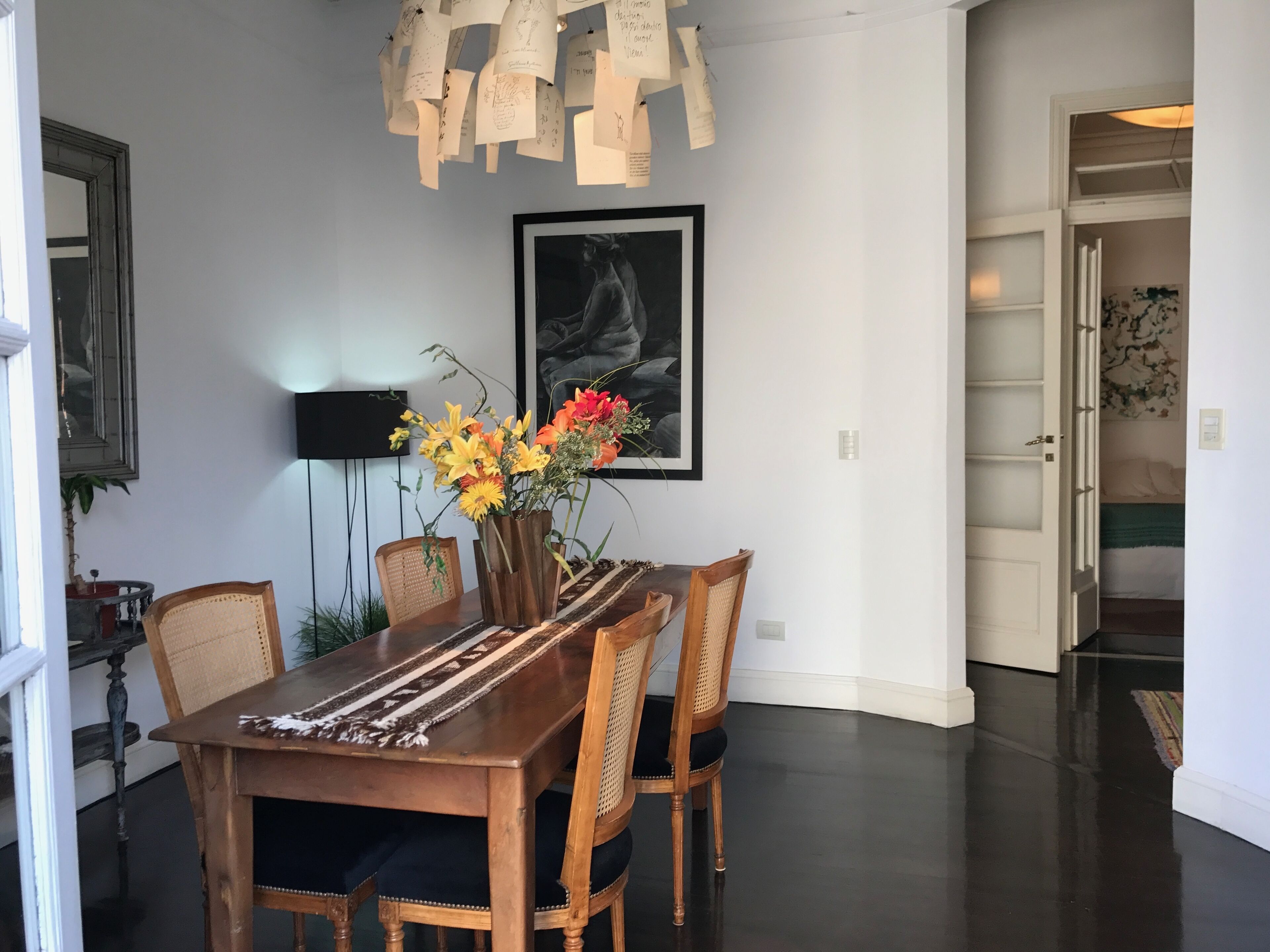 A dining area with a wooden table, chairs, a vase of flowers, a framed artwork, and a wall-mounted light fixture.