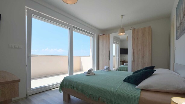 Een slaapkamer met een bed, een houten bankje, een groene deken en uitzicht op de lucht door een groot schuifdeur.