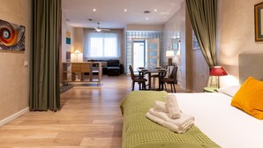 Une chambre d’hôtel avec un lit, un canapé vert, une table à manger avec des chaises, et un coin cuisine à l’arrière-plan.