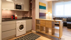 Une cuisine moderne équipée d’un lave-linge, d’un four à micro-ondes et d’un évier. On y trouve un bureau en bois avec des tiroirs et une lampe. Dans l’espace de vie, on aperçoit un canapé et une fenêtre avec des stores.