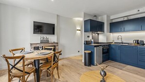 A modern kitchen with blue cabinets, a dining area with a round table and chairs, and a flat-screen TV mounted on the wall. - L’escale 1 - Modern Getaway in the Heart of Honfleur (Honfleur)