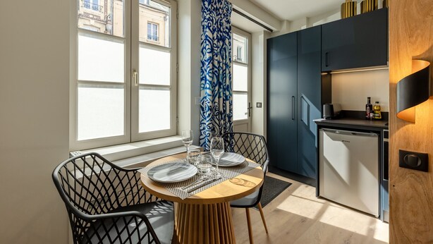 Une cuisine compacte avec un coin repas, comprenant un petit réfrigérateur, une fenêtre donnant sur des immeubles et un rideau à motifs.