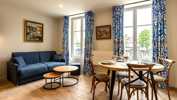 A living room with a blue sofa, a round wooden table, and a dining area with wooden chairs. - L'Escale du Vieux Port 4P - Closed Garage - Sauna - Air Conditioning (Honfleur)