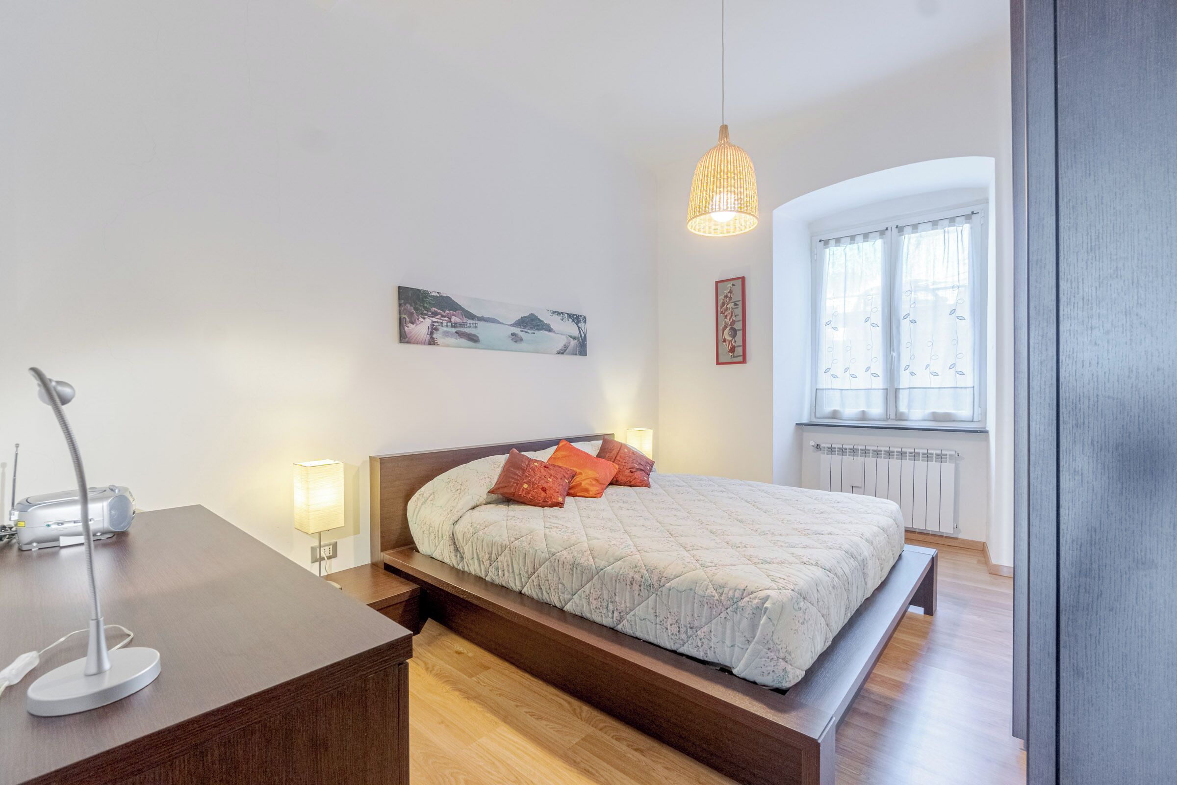 Un dormitorio con cama, lámparas de noche, escritorio con lámpara, un cuadro en la pared y una ventana con cortinas.