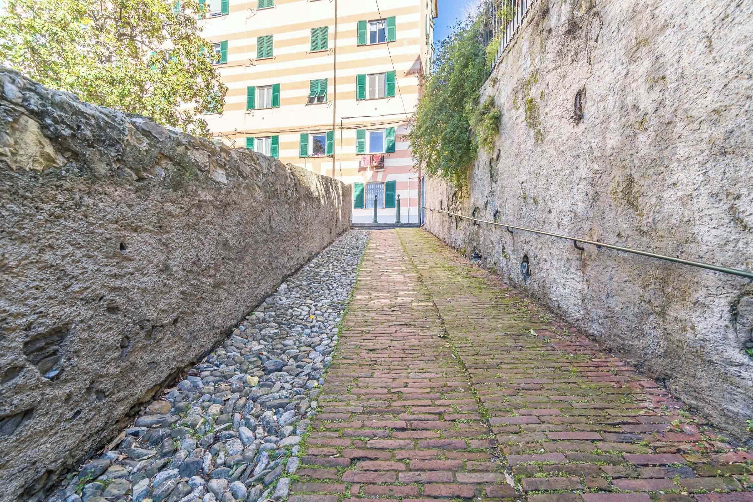 A narrow cobblestone alleyway flanked by stone walls, leading to a building with striped windows and a balcony.