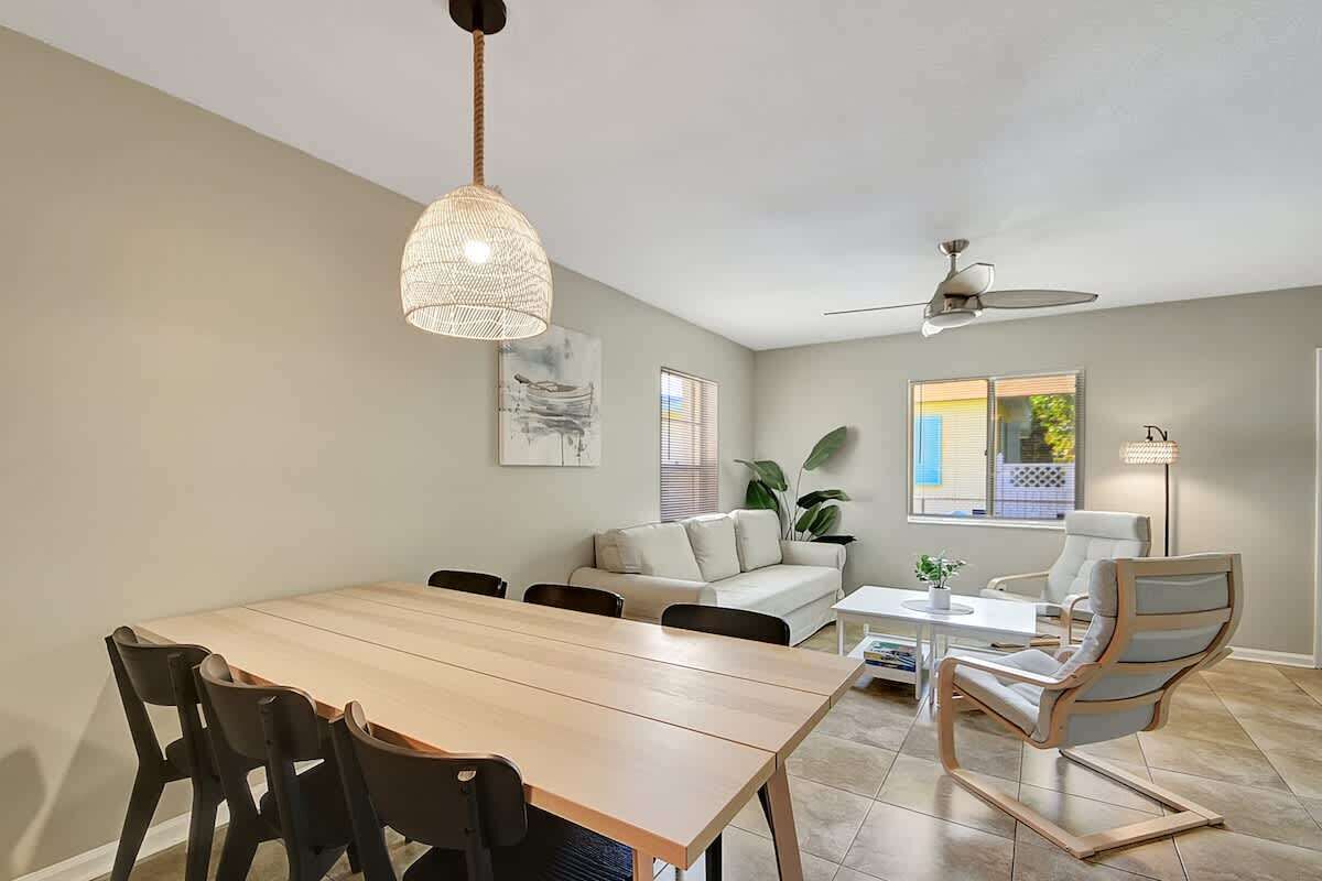 Un coin repas comprenant une table en bois, des chaises, un luminaire suspendu, un ventilateur de plafond, un canapé et une fenêtre avec des stores.