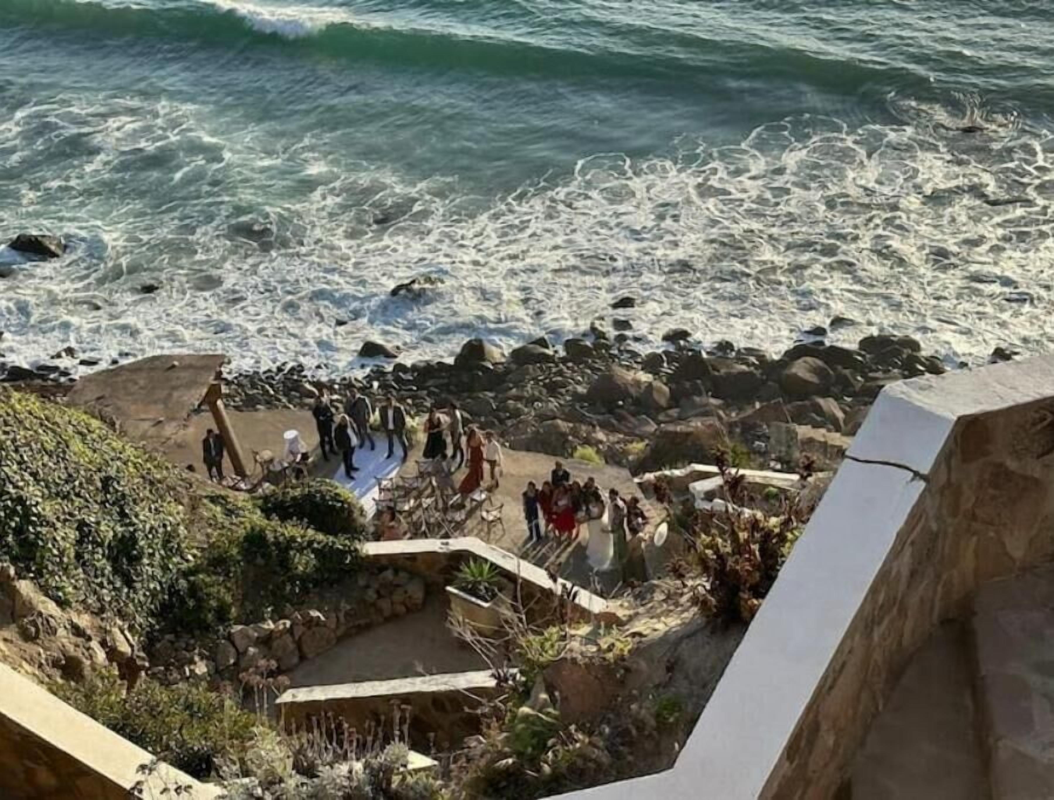 Ett kustområde med en stenig strandlinje, en grupp människor samlade nära vattnet och en stentrappa som leder ner till stranden.