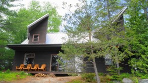 Une cabane avec un toit en pente, une véranda avec des chaises en bois et une forêt environnante.