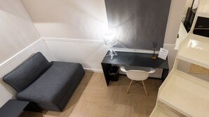 A compact room with a grey sofa, a small desk with a lamp, a white chair, and a white cabinet. - Five Boutique Rooms (Naples)