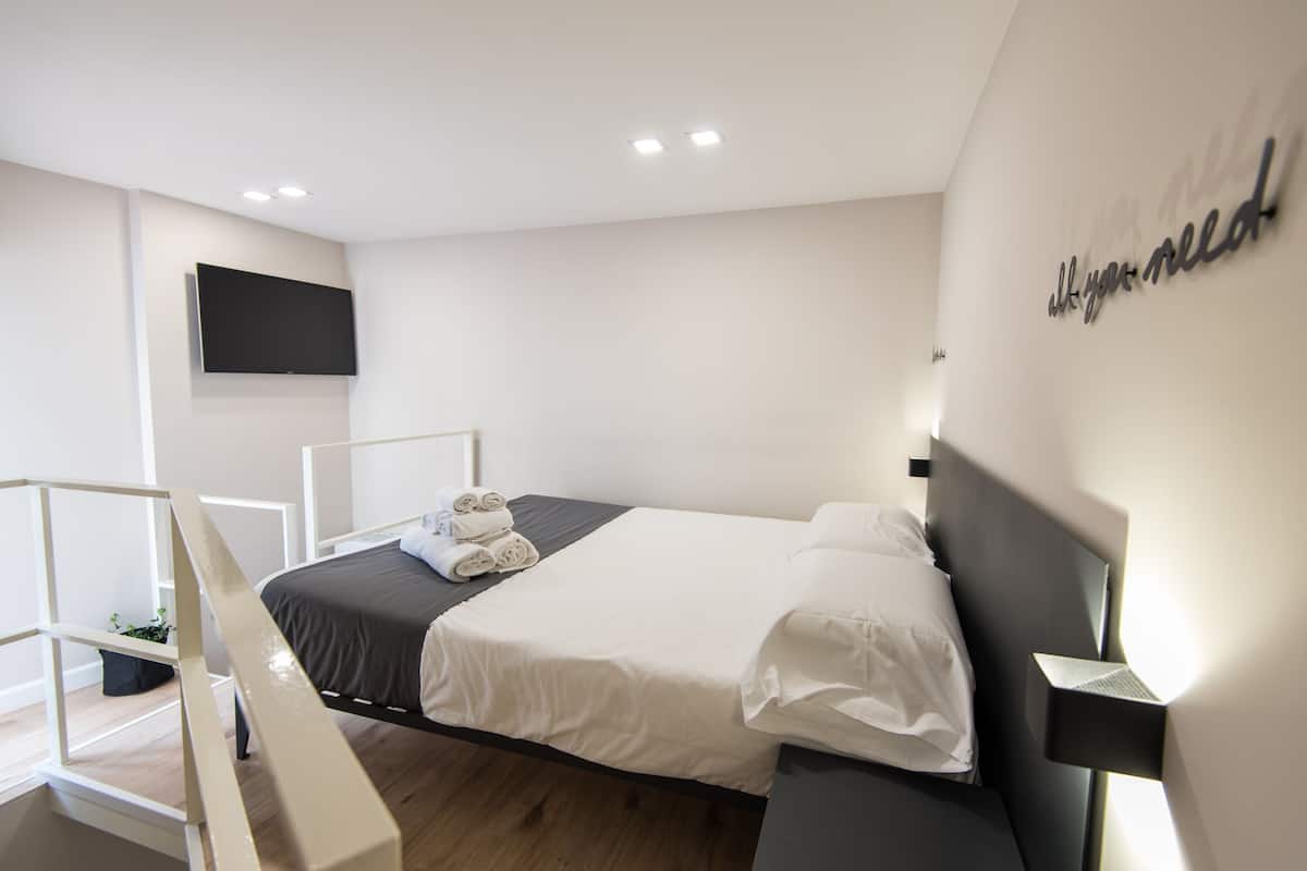 Une chambre d’hôtel moderne équipée d’un lit, d’un téléviseur à écran plat fixé au mur et d’une table de chevet avec une lampe.