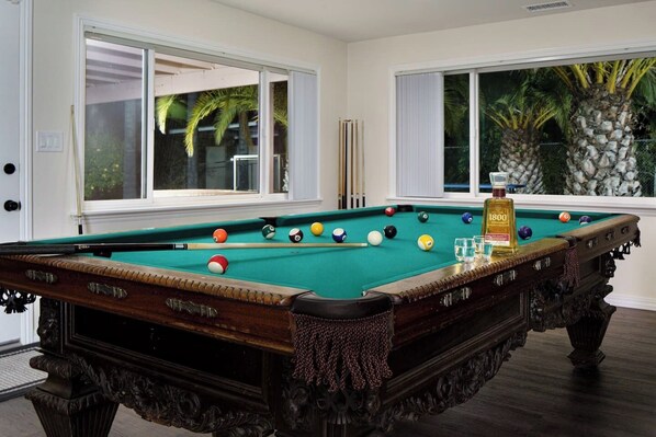 Une table de billard avec des billes et des queues, une bouteille de spiritueux et une fenêtre donnant sur des palmiers.