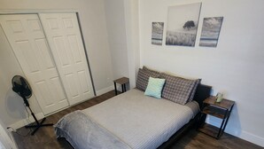Une chambre avec un grand lit, un plancher en bois, un ventilateur sur pied et une petite table avec une bougie.