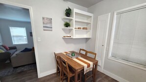 Un coin repas avec une table et des chaises en bois, un mur blanc avec des étagères, une fenêtre avec des stores et une porte.