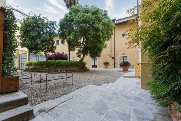 A courtyard with stone flooring, a metal bench, potted plants, and a building with a yellow facade. - Apartments Florence- Dragomanni Loft (two) with garden (Firenze)