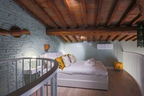 Quarto com cama branca, vigas de madeira, parede de tijolos e um banquinho amarelo.
