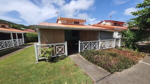 A small, single-story building with a red roof and white exterior walls. It features a covered entrance with a white railing and a small garden area with various plants.