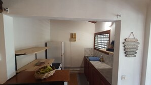 A small kitchen with a refrigerator, a window with bars, a wooden shelf, a basket of bananas, and a wall-mounted sign with French text. - Bungalow Style Studio vue Mer, Accès Direct Plage Véhicule de Location Possible (La Trinité)