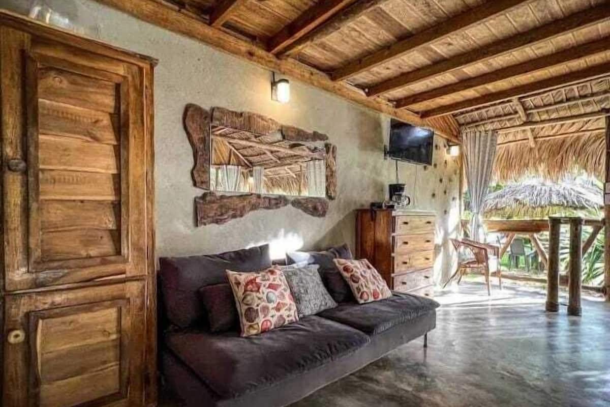 Une pièce avec un plafond en bois, un canapé agrémenté de coussins à motifs, un téléviseur fixé au mur et une structure de toit en chaume.