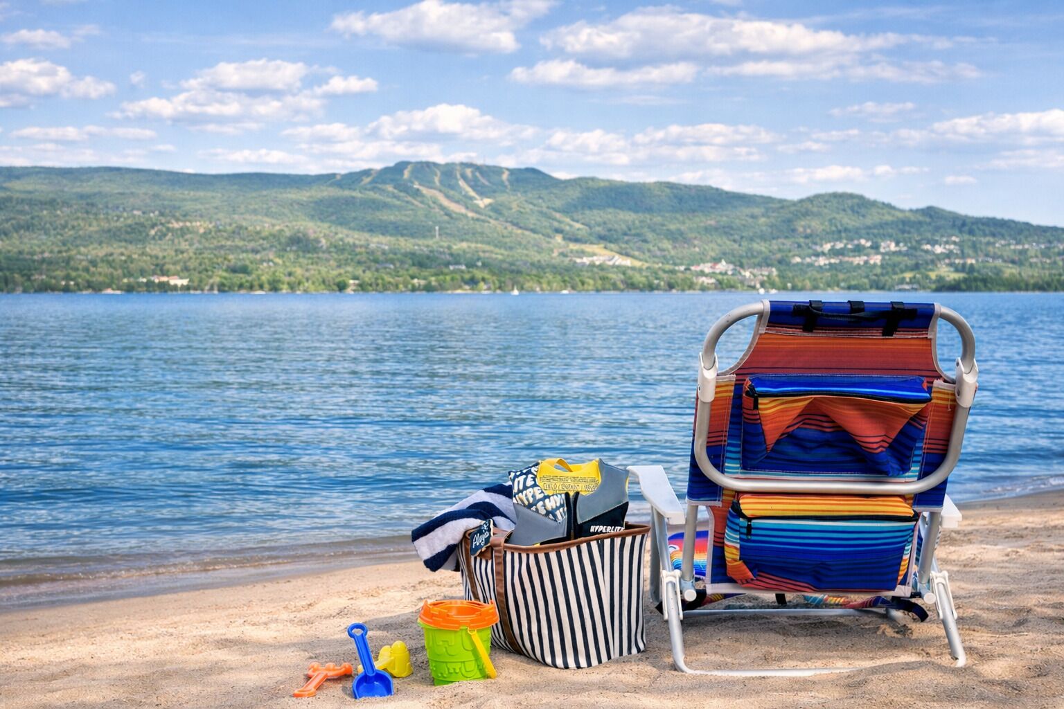 A beach chair and a bag with toys on a sandy shore, overlooking a lake with hills in the background.