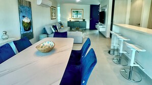 A modern living room with a dining area, featuring a large table, blue chairs, a sofa, and a kitchen in the background. - NEW 3/2 POOLFRONT. FULL BACKUP GENERATOR, 2 POOLS, NEAR BEACH, GYM, 2 PARKINGS. (Carolina)