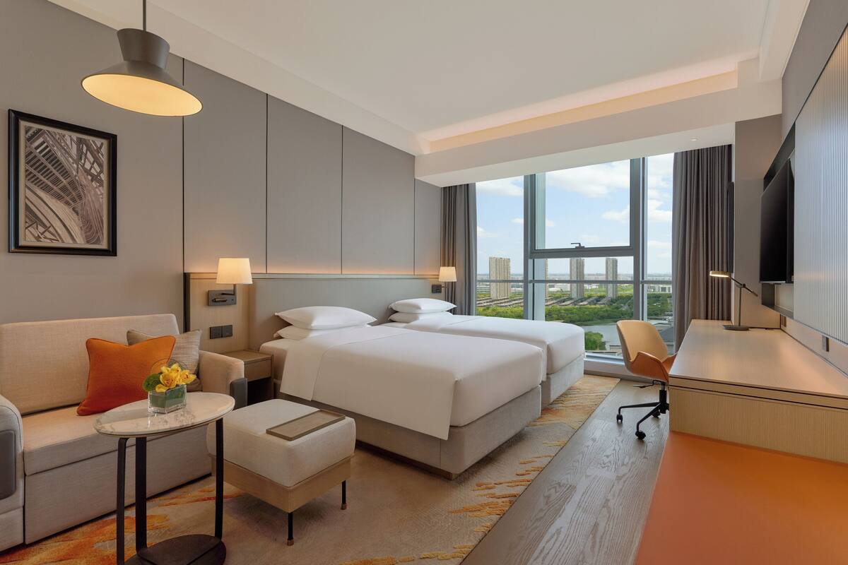 A modern hotel room with a large bed, a desk, a sofa, and a view of the city.