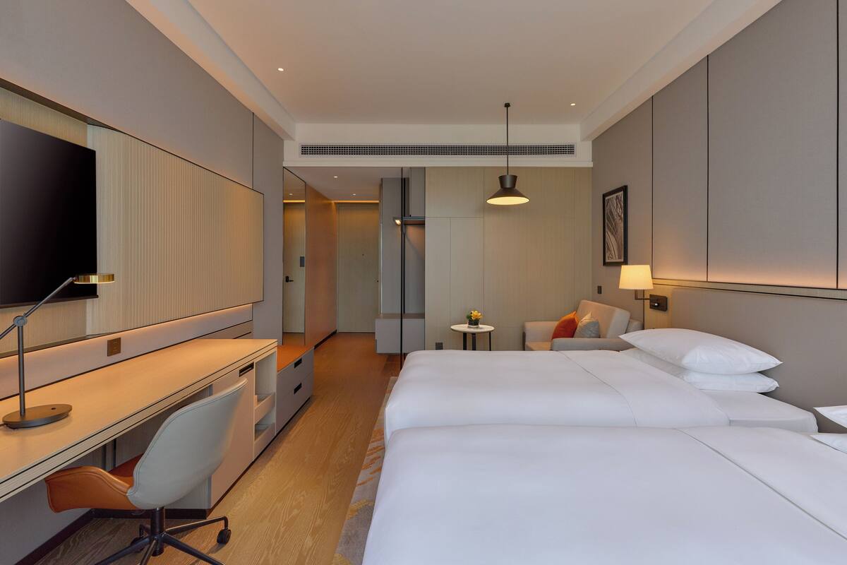 A hotel room with a large bed, a desk with a chair, a small table with a lamp, and a television on the wall.