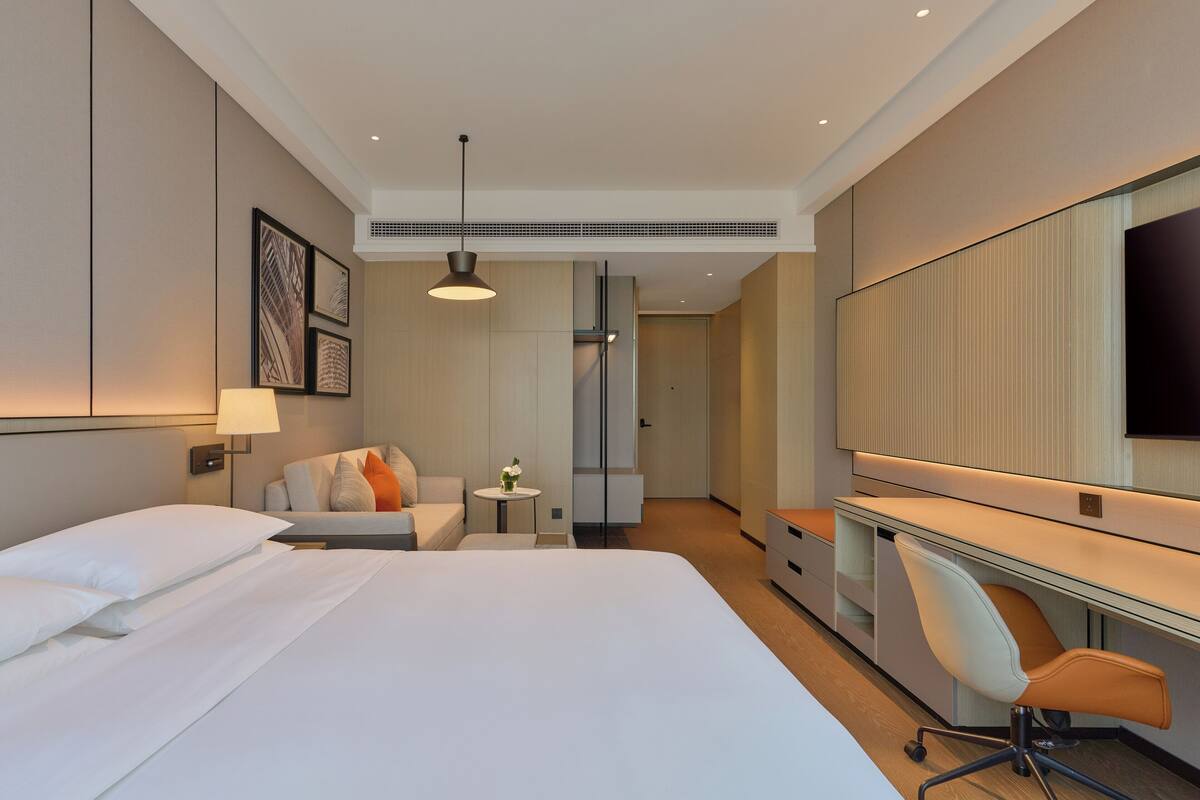 A modern hotel room with a large bed, a desk with a chair, a TV, and a small table with a lamp.