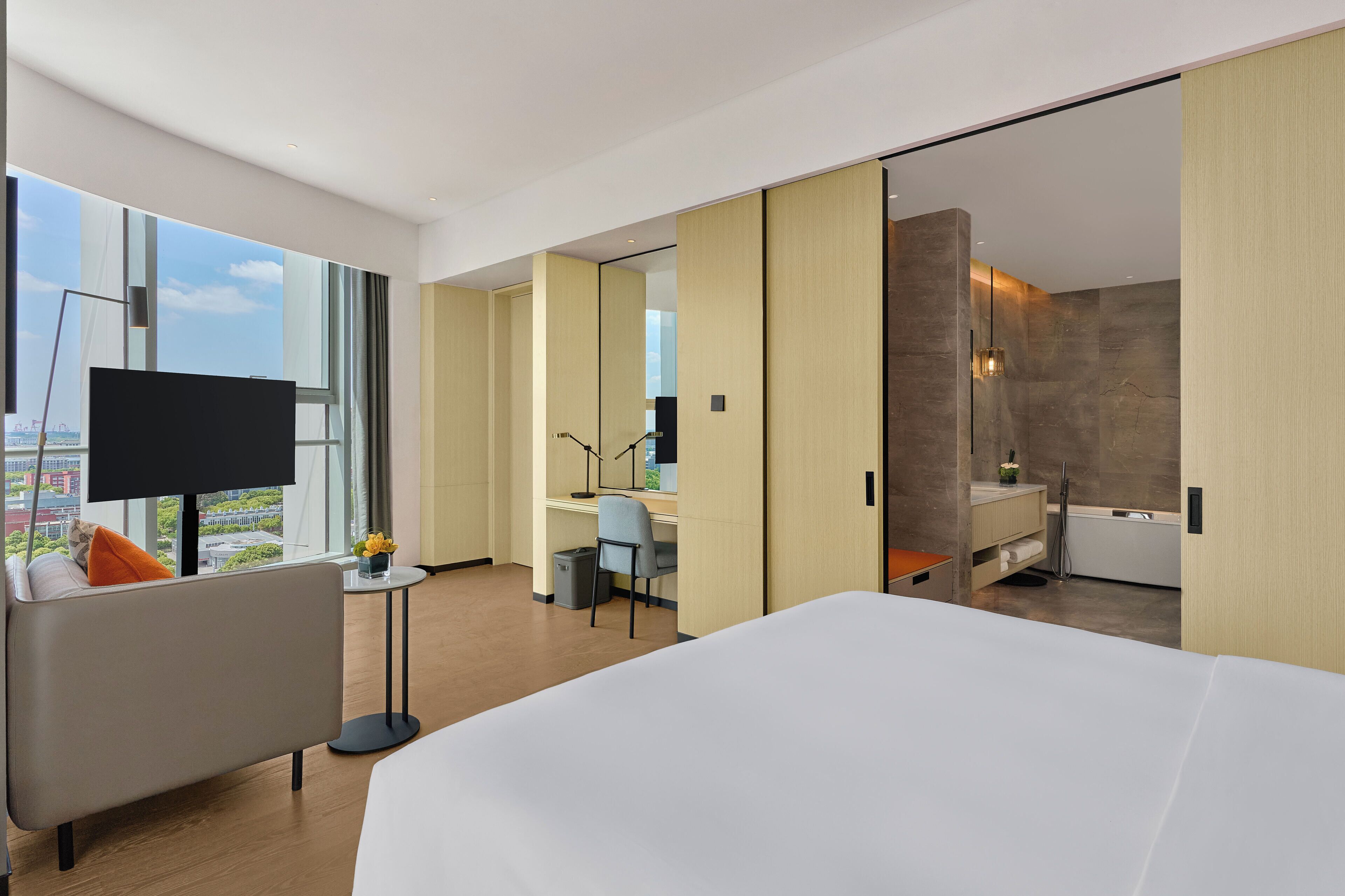 A modern hotel room with a large bed, a desk with a chair, a TV, and a bathroom with a bathtub.