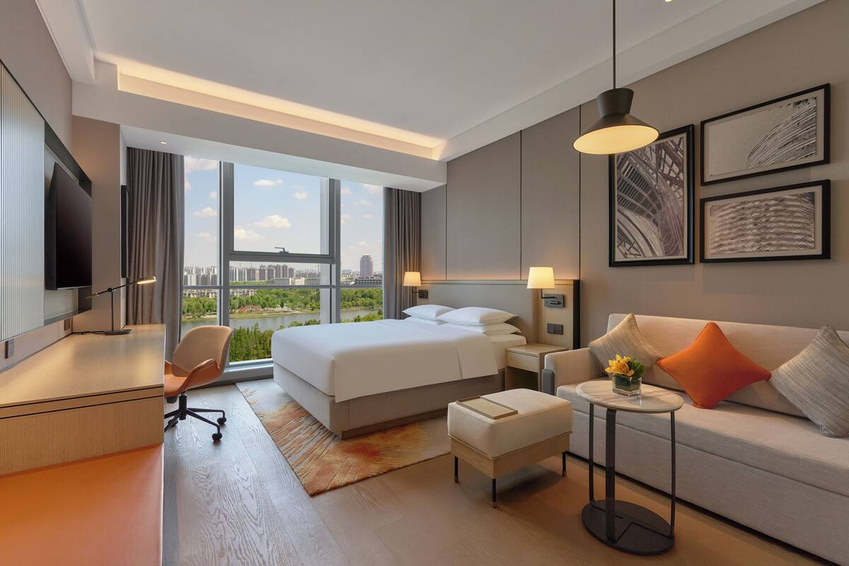 A modern hotel room with a large bed, a desk, a sofa, and a view of the city.