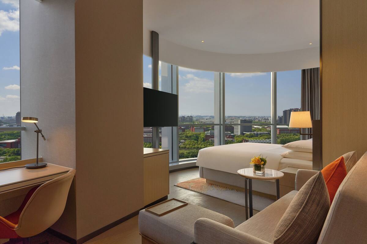 A modern hotel room with a large bed, a desk, a chair, a small table, and a TV. The room has large windows offering a city view.