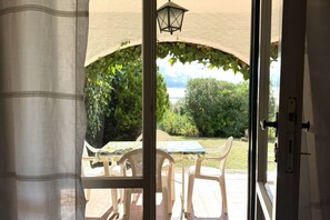 Een open deur die leidt naar een terras buiten met een tafel en stoelen, uitzicht op groen en een lamp.