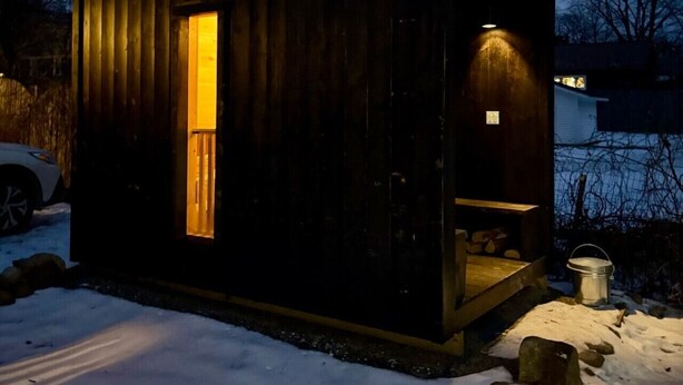 Pieni puinen mökki, jonka sisällä on valot, siinä on savupiippu ja sen ympärillä oleva maa on lumen peitossa.