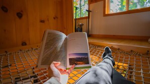 Una persona recostada en una hamaca dentro de una cabaña de madera, con un libro abierto frente a ella.