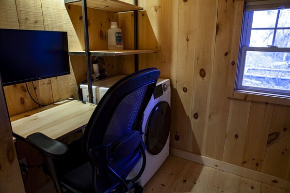 Une pièce compacte comprenant un bureau, un lave-linge, une télévision et une fenêtre donnant sur un paysage agréable.