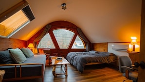 Ein gemütliches Schlafzimmer im Dachgeschoss mit einem Schlafplatz auf Hochbett, einem kleinen Tisch, einem Stuhl und einer Couch.