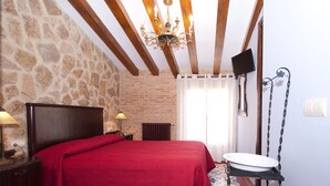 A bedroom with a stone wall, wooden beams, a red bedspread, a bedside table with a lamp, a radiator, and a bathroom with a sink and mirror. - Do you need to take a break between mountains in a charming village in Alicante? (La Torre de les Maçanes)