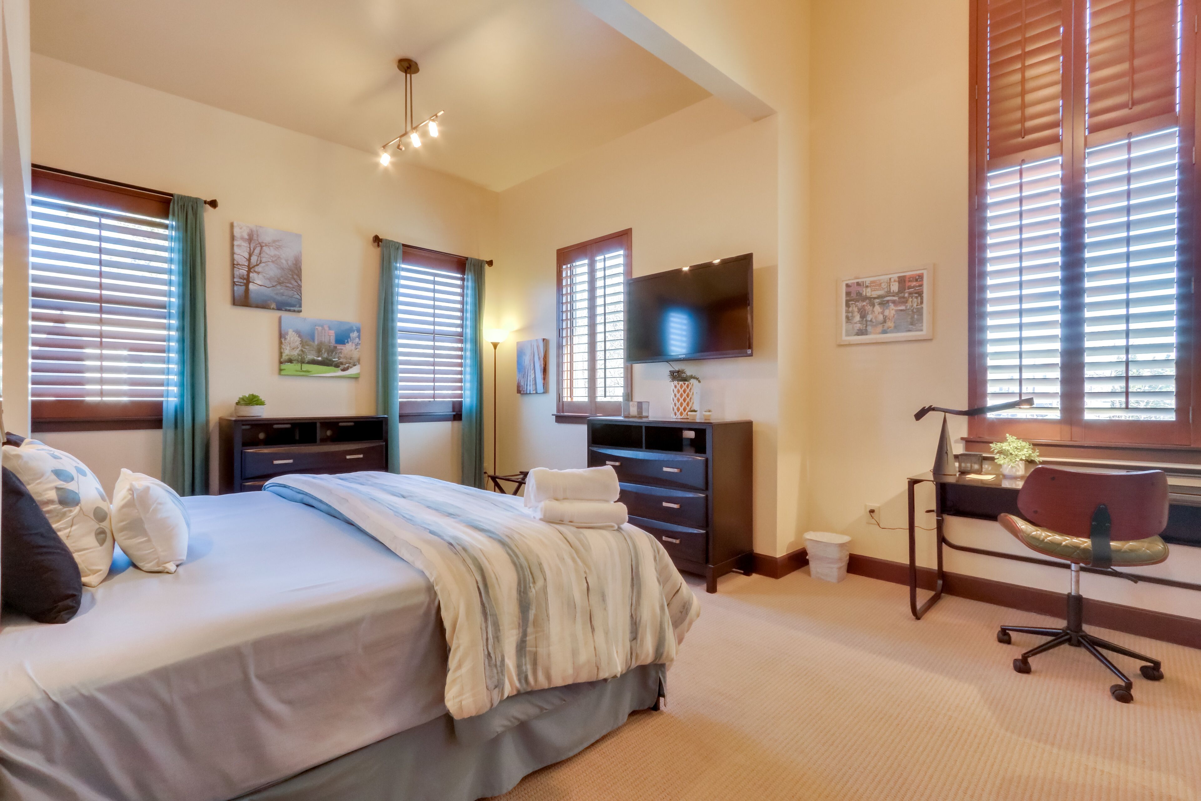 A hotel room with a bed, a desk, a chair, a TV, and large windows with blinds.