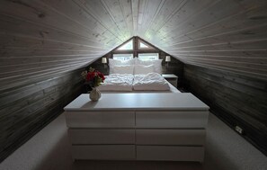 Un dormitorio bajo del ático, pequeño y estrecho, con techo inclinado, cama con ropa blanca, una mesita de noche con un jarrón de flores y una ventana al fondo.