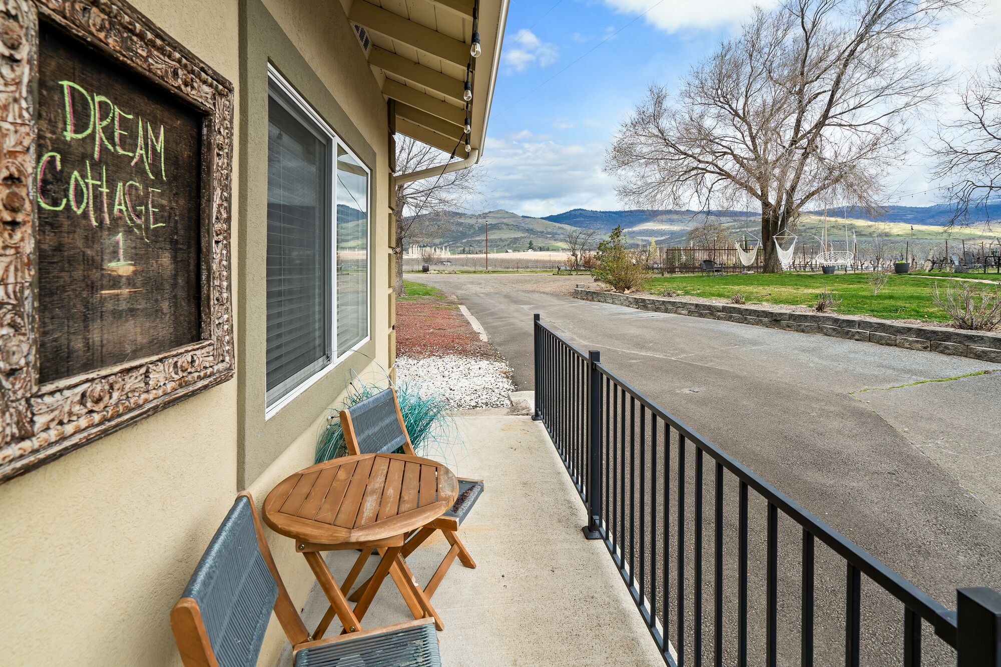 A cozy outdoor seating area with a wooden table and chairs, a chalkboard sign reading "Dream Cottage," and a view of a rural landscape with hills and a tree.