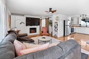 A modern living room with a grey sofa, a fireplace, a television, and a kitchen area with white cabinets and stainless steel appliances.