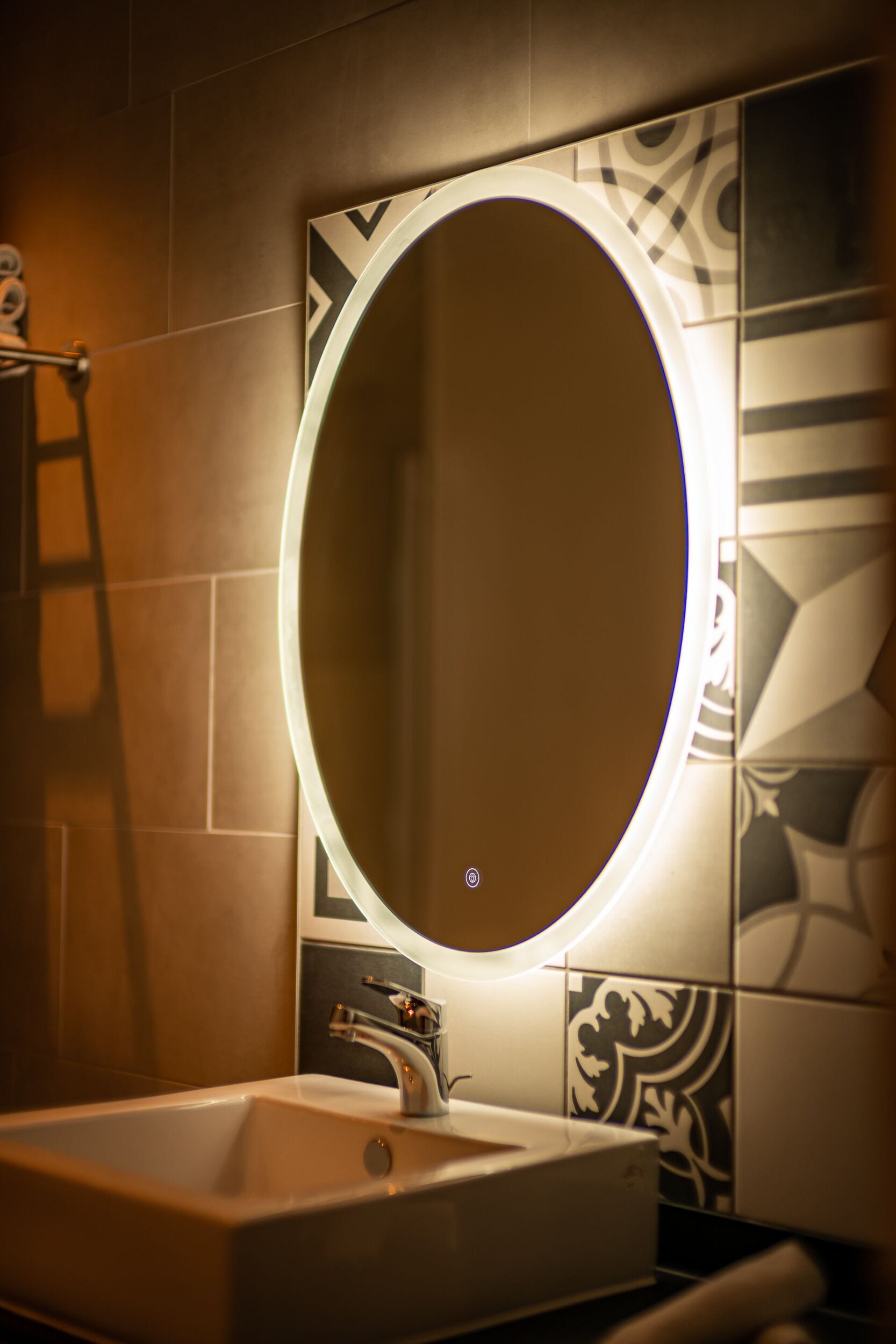 Une salle de bain avec un lavabo rectangulaire, un robinet moderne et un miroir fixé au mur, surmonté d’un luminaire circulaire.