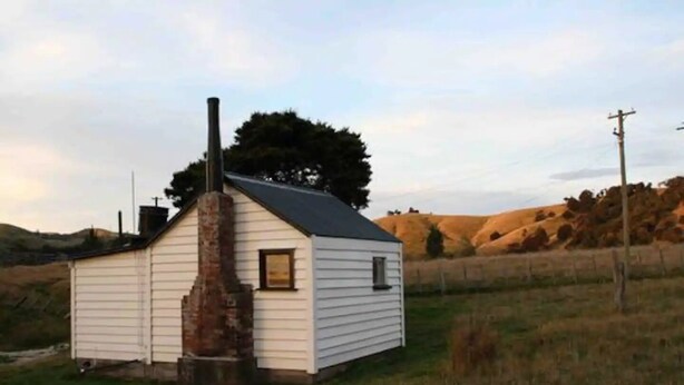 A small, single-story cabin with a chimney, surrounded by a grassy field and hills in the background. - Walnut Whare (Te Whiti)