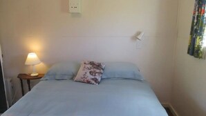 A bedroom with a bed, a nightstand with a lamp, and a power outlet on the wall. - Walnut Whare (Te Whiti)