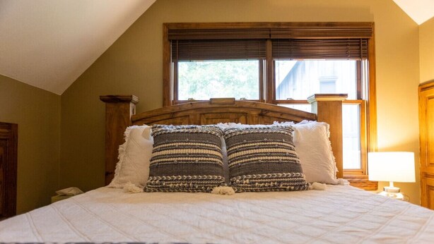 A neatly made bed with a patterned bedspread and two decorative pillows, a wooden headboard, and a window with blinds.
