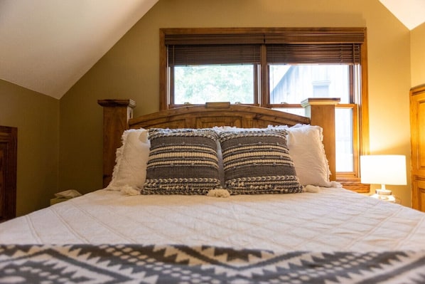 A neatly made bed with a patterned bedspread and two decorative pillows, a wooden headboard, and a window with blinds.