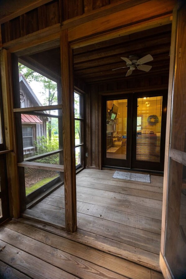 Eine Holzbalkon mit Deckenventilator, Blick auf ein Gebäude mit rotem Dach und eine Tür zu einem Innenraum mit Jalousien.