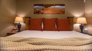 Una cama bien hecha con un edredón texturizado, dos lámparas de noche y tres cuadros en la pared que representan paisajes desérticos.