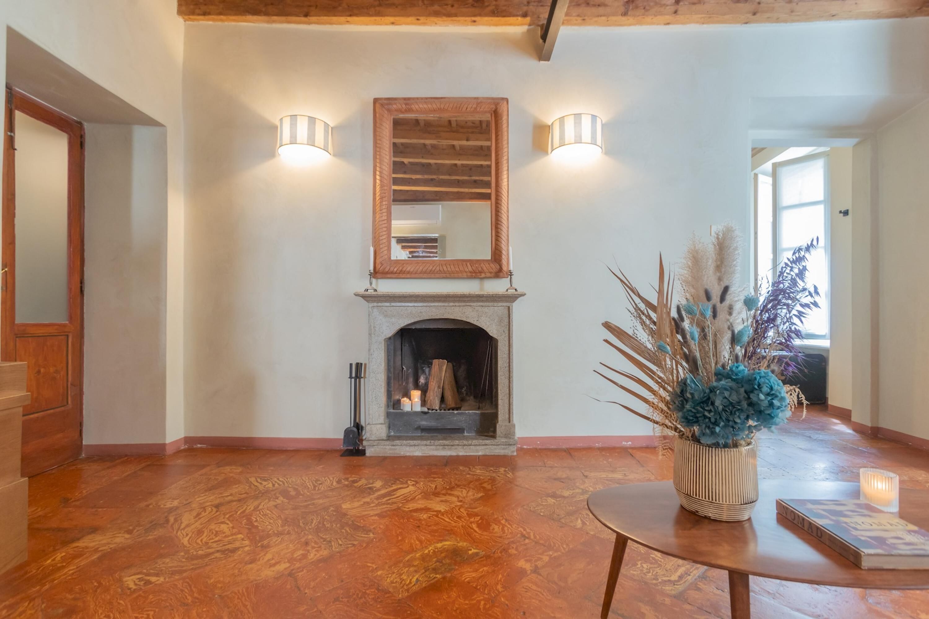 A spacious room with a fireplace, wooden flooring, a round table with a book and candle, and a vase with dried plants.