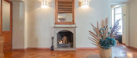 A spacious room with a fireplace, wooden flooring, a round table with a book and candle, and a vase with dried plants.