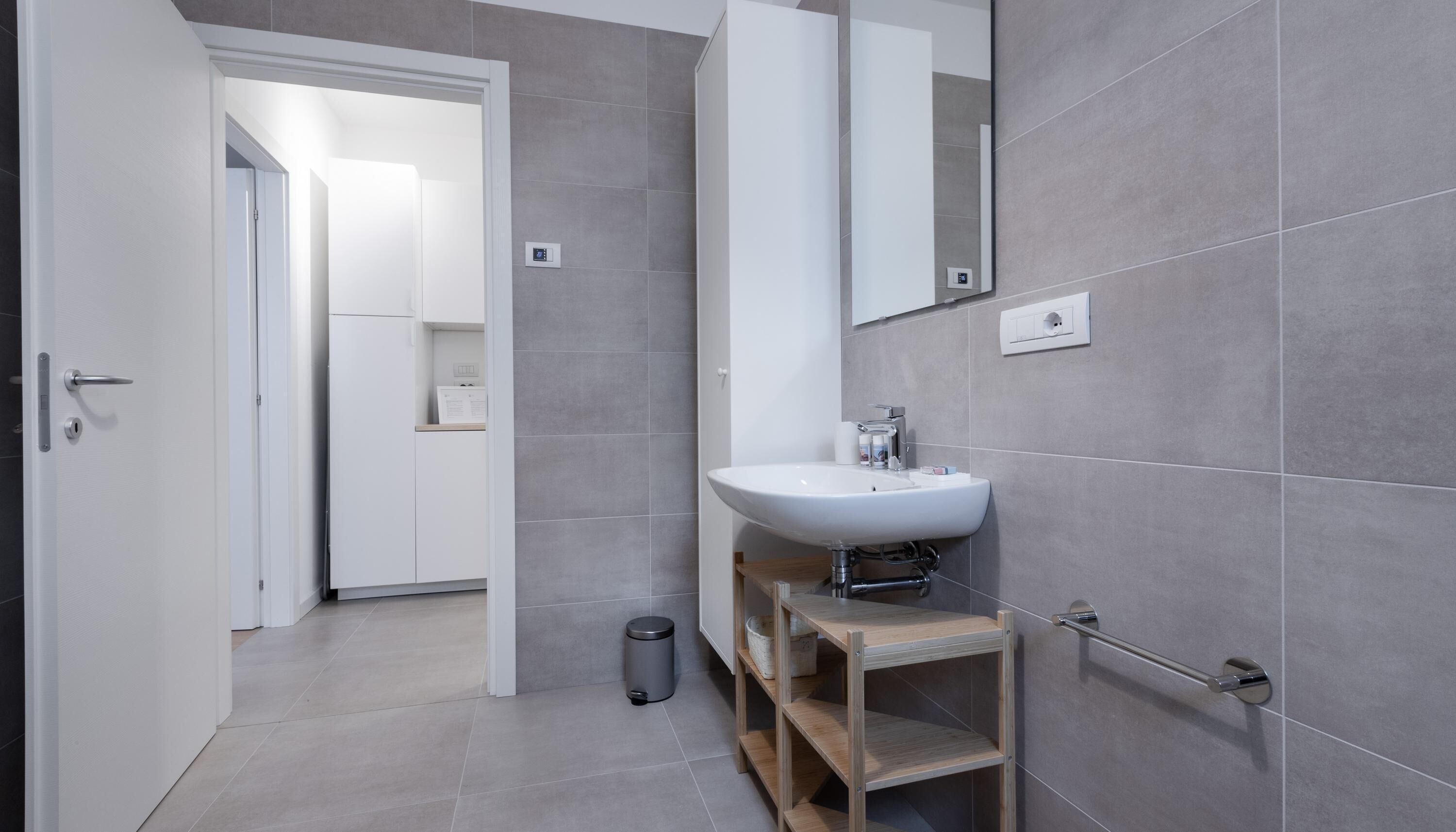 A modern bathroom with a white sink, a mirror, and grey tiled walls. There is a trash bin and a wooden shelf with toiletries.